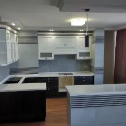 Nowoczesna kuchnia z ciemnymi dolnymi szafkami i białymi górnymi, jasny blat, płytki z poziomymi paskami, oświetlenie sufitowe i wiszące lampy.