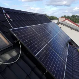 Panele fotowoltaiczne zamontowane na dachu z czarnej dachówki, widoczny system zabezpieczeń linowych oraz okno dachowe.