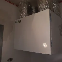 Biała jednostka rekuperacyjna marki WANAS zamontowana pod sufitem, z widocznymi, owiniętymi folią aluminiową przewodami wentylacyjnymi. Ściana w stanie surowym, z widocznym otworem instalacyjnym.
