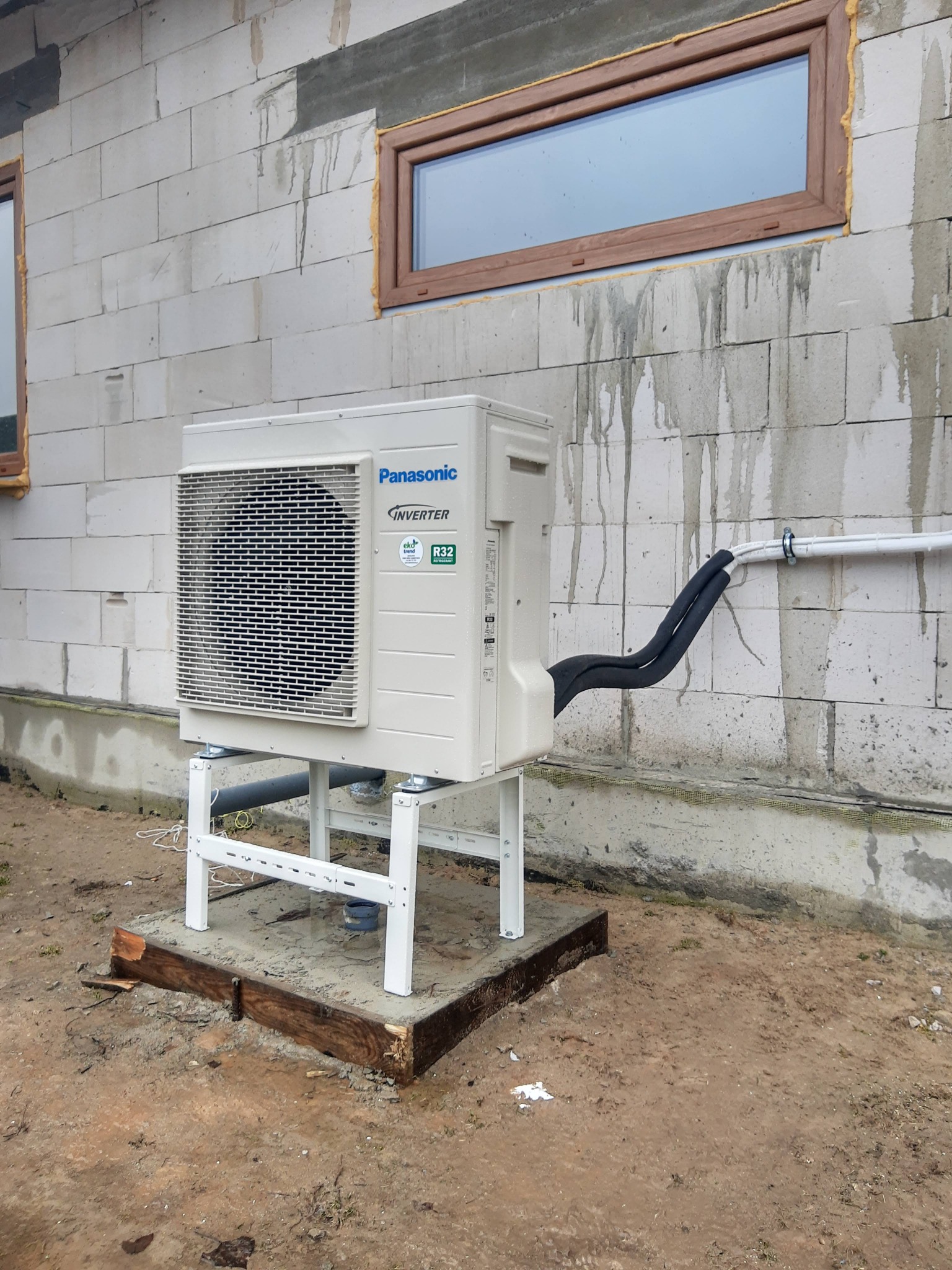 Zewnętrzna jednostka klimatyzacji Panasonic Inverter zamontowana na białym stelażu, umieszczona na betonowej podstawie obłożonej drewnem, przy ścianie budynku z białych bloczków z widocznymi...