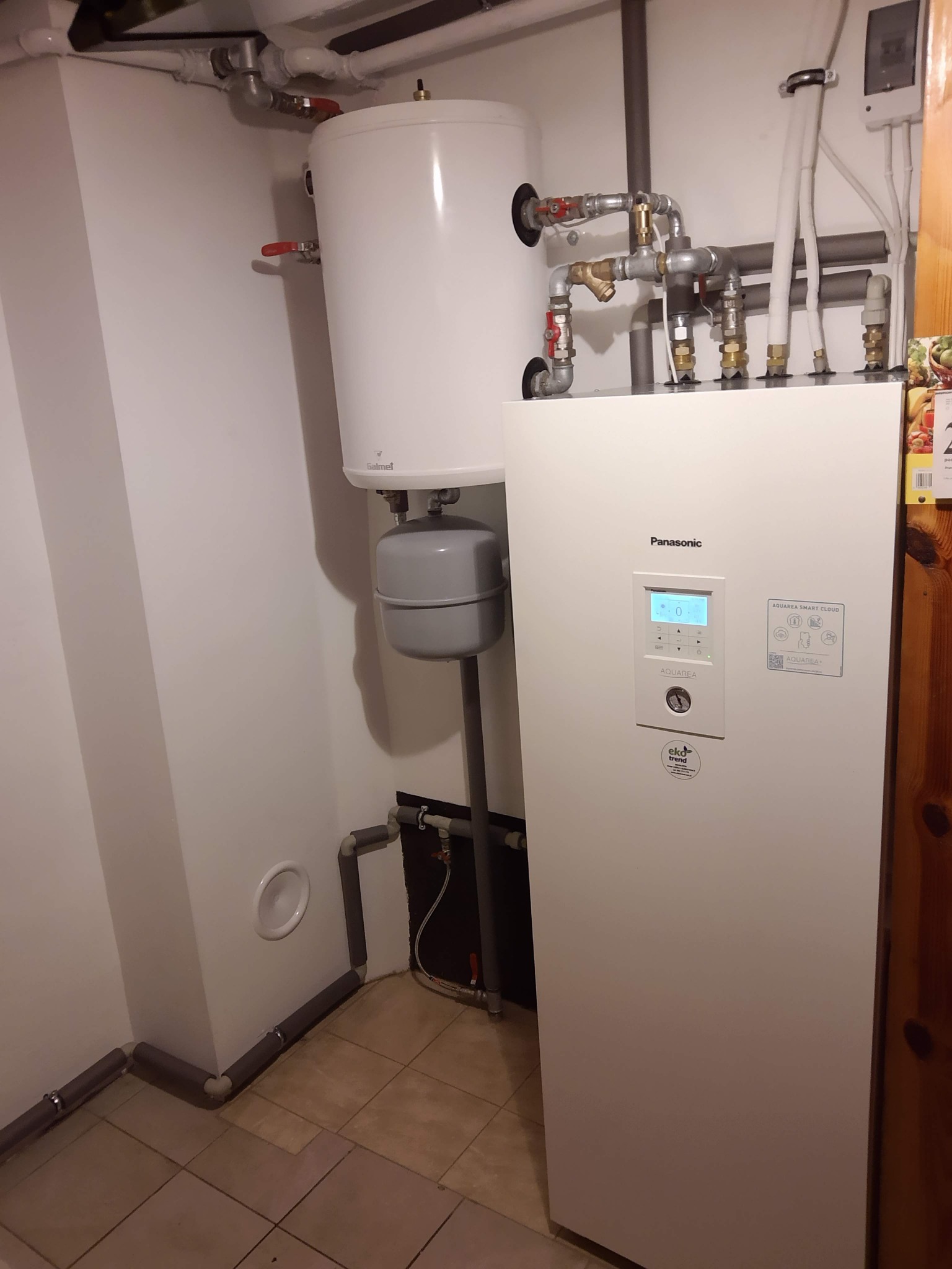 Biała pompa ciepła Panasonic Aquarea w kotłowni, widoczne podłączenia hydrauliczne i zasobnik ciepłej wody Galmet.