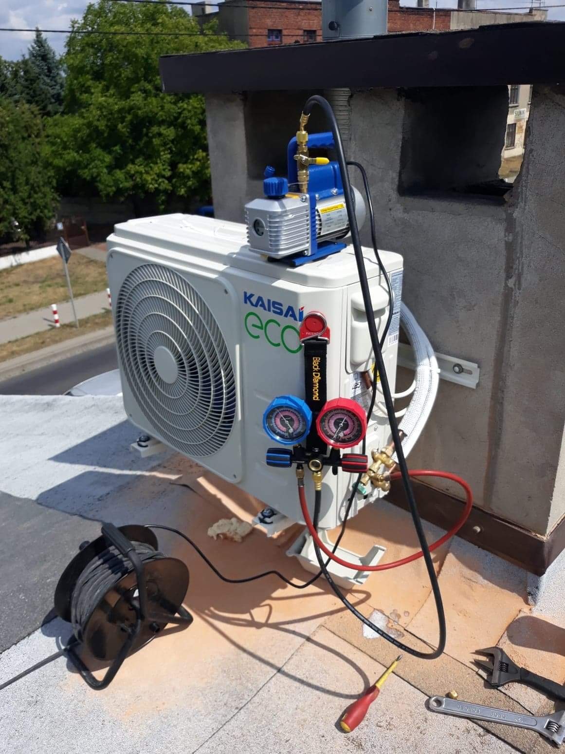Biała jednostka zewnętrzna klimatyzacji Kaisai Eco zamontowana na dachu budynku, podłączona do pompy próżniowej i manometrów, narzędzia leżą obok.