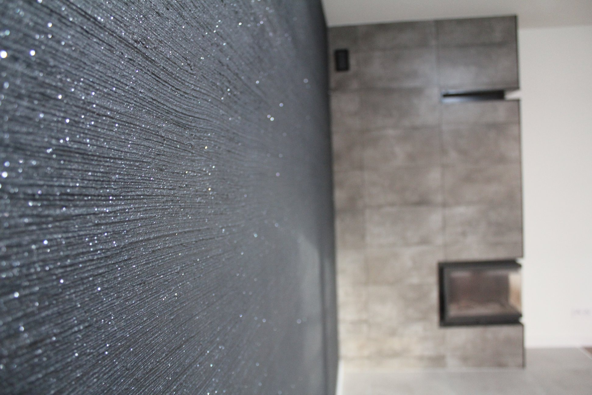 Czarna ściana z dekoracyjną fakturą i srebrnymi drobinkami, w tle fragment obudowy kominka z szarych płyt.