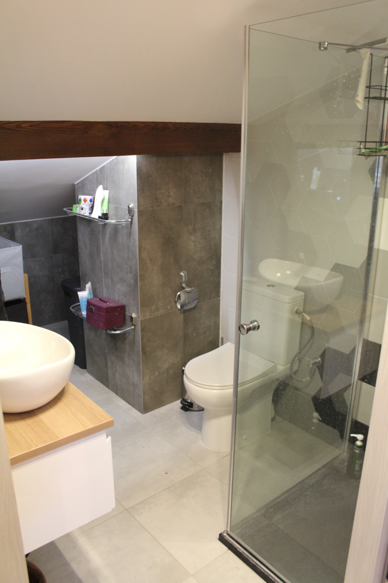 Nowoczesna łazienka na poddaszu z drewnianą belką stropową, szarą glazurą, kabiną prysznicową ze wzorem, umywalką nablatową na drewnianym blacie i toaletą.