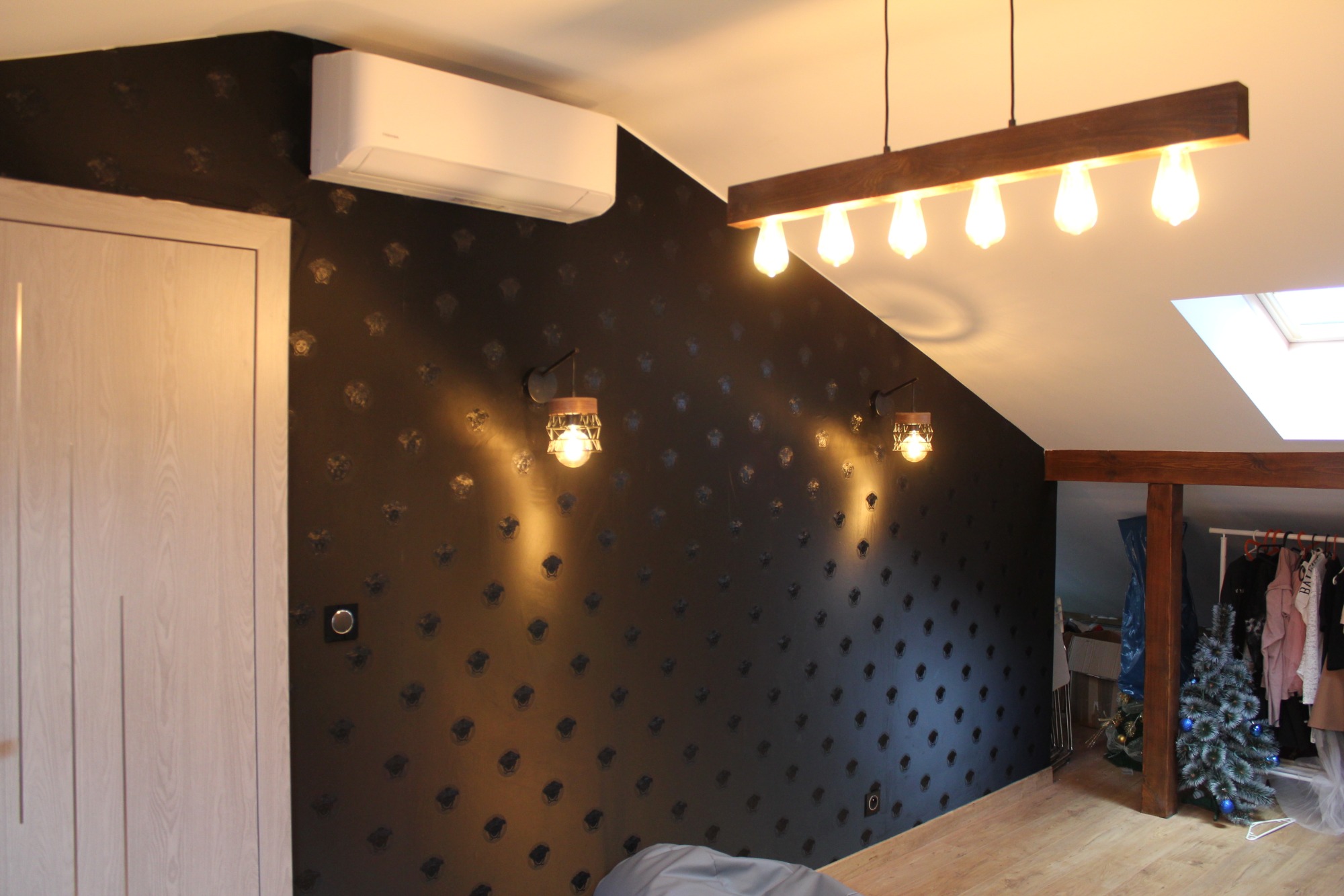 Pokój na poddaszu z czarną ścianą w złote wzory, oświetlony dekoracyjnymi lampami ściennymi i wiszącą lampą z drewnianą belką, widoczny klimatyzator i fragment garderoby z choinką.