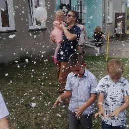 Dzieci bawiące się w pianie na zewnątrz budynku, mężczyzna trzyma dziewczynkę z balonem w ręku.