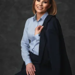 Portret uśmiechniętej kobiety w błękitnej koszuli i granatowym garniturze, siedzącej na krześle na tle ciemnoszarej, teksturowanej ściany. Marynarka nonszalancko zarzucona na ramię, złoty...