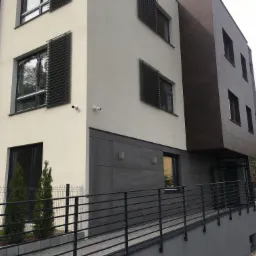 Nowoczesny budynek z elewacją w odcieniach bieli, szarości i brązu, z poziomymi żaluzjami okiennymi i rampą dla wózków inwalidzkich.