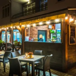 Oświetlony ogródek letni z drewnianym barem, stolikami z kwiatami i kelnerką w tle, przytulna atmosfera.