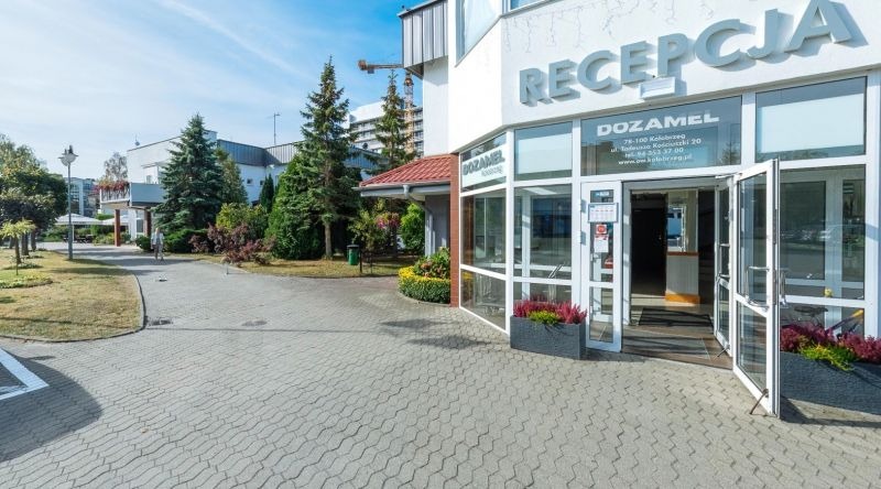 Wejście do recepcji hotelu Dozamel w Kołobrzegu, widok z zewnątrz na otwarte drzwi i chodnik z kostki brukowej.