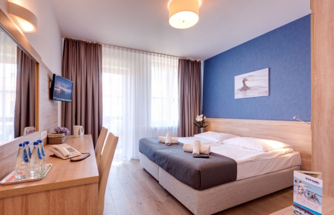 Jasny pokój hotelowy z łóżkiem typu king-size, biurkiem z butelkami wody i telewizorem, ściana akcentująca w kolorze niebieskim z obrazem ptaka