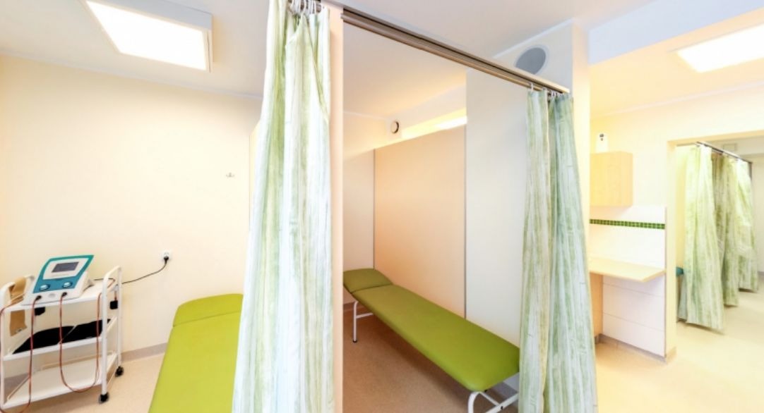 Gabinet rehabilitacyjny z zielonymi leżankami, zasłonami w pastelowe wzory i aparaturą do fizykoterapii na wózku.