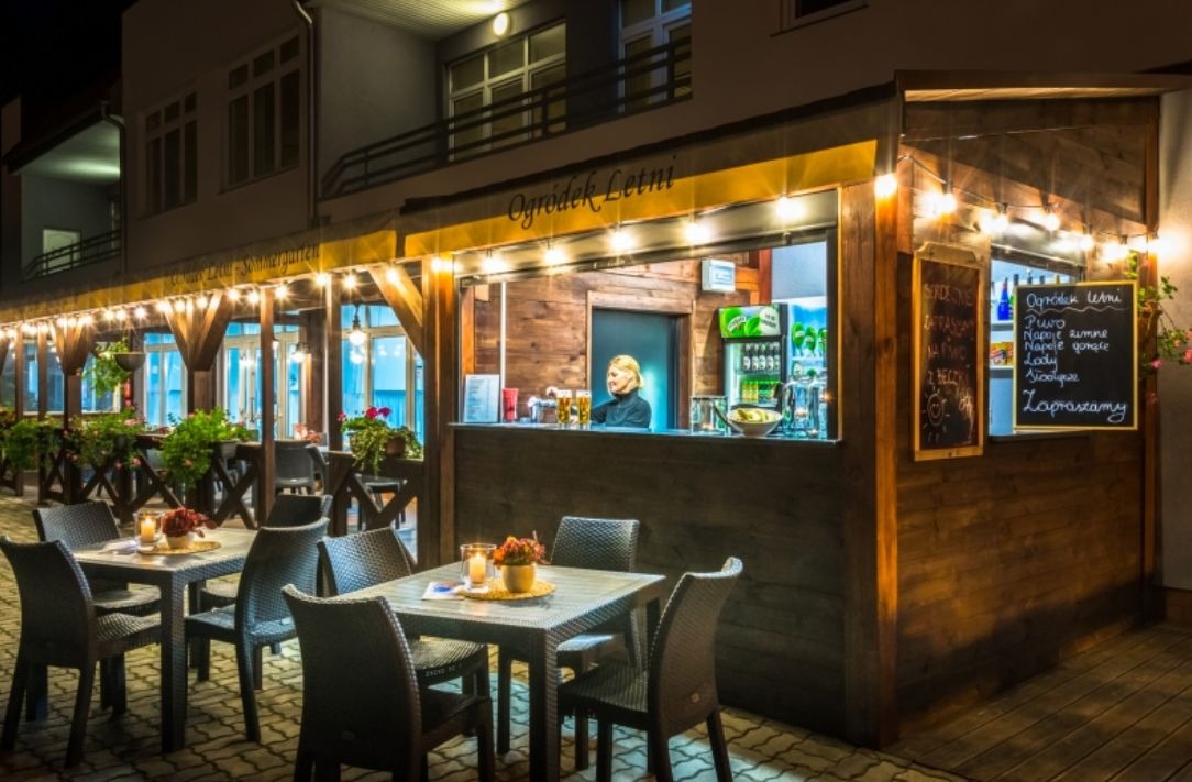 Oświetlony ogródek letni z drewnianym barem, stolikami z kwiatami i kelnerką w tle, przytulna atmosfera.