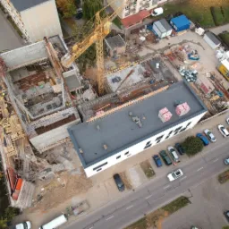 Widok z drona na plac budowy z żółtym dźwigiem, widoczne szalunki, zbrojenia i betonowe elementy konstrukcyjne w trakcie wznoszenia budynku, na zewnątrz zaparkowane samochody i kontenery budowlane.