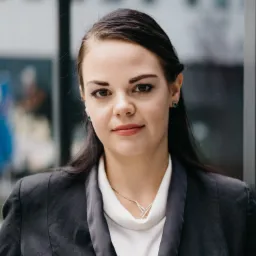 Portret kobiety w ciemnej marynarce i białej bluzce, z delikatnym naszyjnikiem i małymi, zielonymi kolczykami, pozującej na tle szklanej ściany.