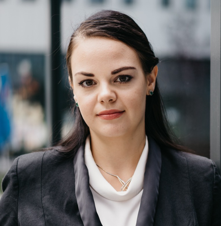 Portret kobiety w ciemnej marynarce i białej bluzce, z delikatnym naszyjnikiem i małymi, zielonymi kolczykami, pozującej na tle szklanej ściany.