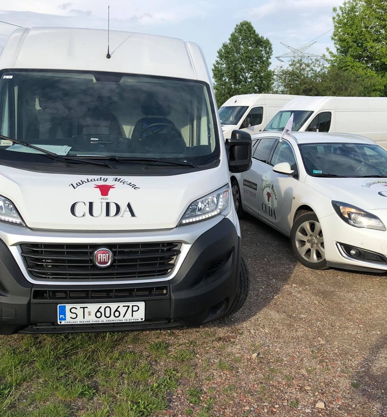Flota pojazdów firmowych Zakłady Mięsne CUDA: biały Fiat Ducato z logo firmy na masce, obok białe kombi Opel z flagą i logo firmy, w tle kolejne białe busy.