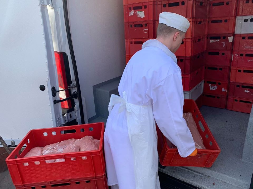 Pracownik w białym uniformie, z widocznymi pomarańczowymi rękawiczkami, przenosi czerwoną skrzynkę z surowym produktem do chłodni samochodowej, w tle ściana ułożona z czerwonych skrzynek.