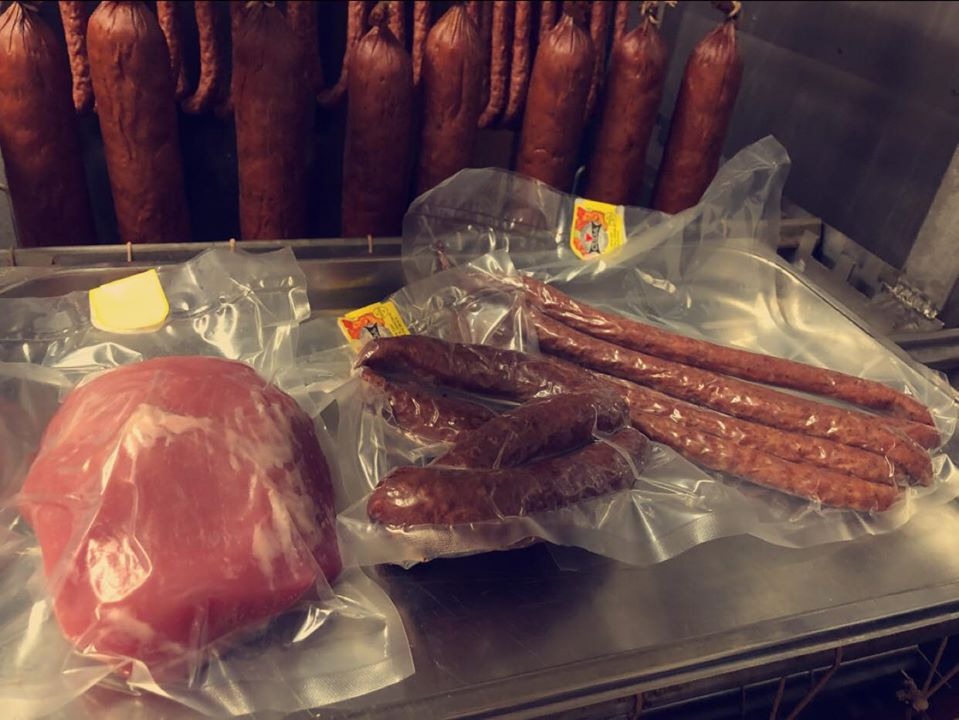 Pakowane w folię wędliny: okrągły kawałek surowego mięsa oraz kilka rodzajów kiełbas na metalowej półce, w tle wiszące wędzonki.