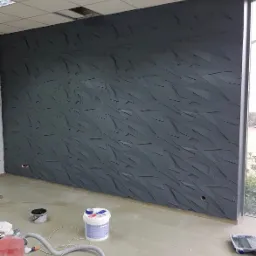 Ściana biurowa z trójwymiarową, geometryczną fakturą w odcieniach szarości, widoczne narzędzia malarskie i wiadro z farbą na podłodze, duże okno po prawej stronie.