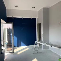 Wnętrze w trakcie odświeżania, ściany w odcieniach szarości i granatu, widoczne narzędzia malarskie, drabina rozstawiona w pomieszczeniu, wpadające promienie słoneczne przez otwarte drzwi balkonowe.