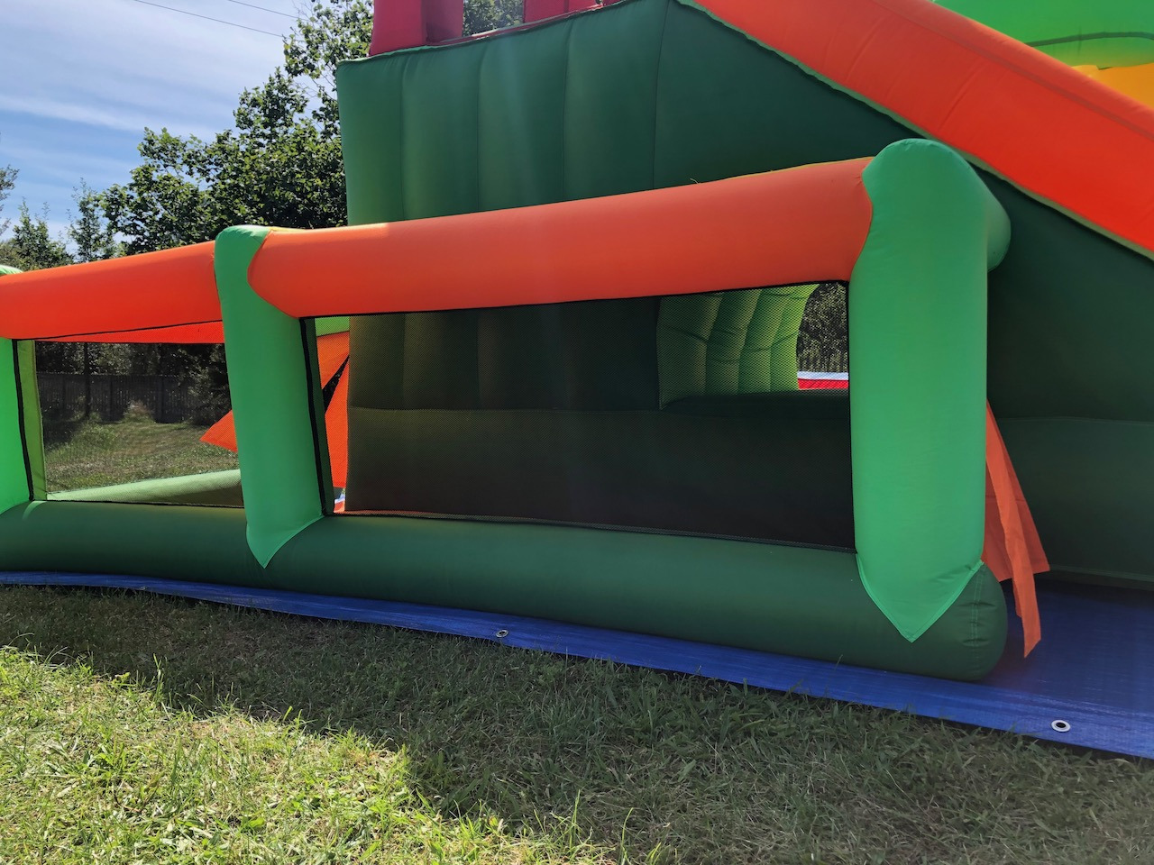 Fragment dmuchanego zamku w odcieniach zieleni i pomarańczy, z widoczną siatką zabezpieczającą i fragmentem niebieskiej maty na trawie.