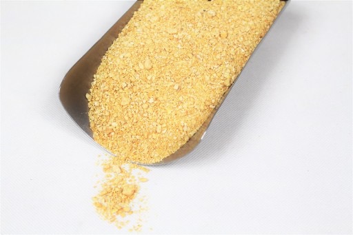 Metalowa łopatka wypełniona drobnymi, jasnobrązowymi granulkami, rozsypującymi się na białe tło.