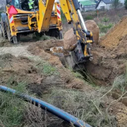 Żółta koparko-ładowarka JCB z operatorem w kamizelce odblaskowej, wykop z ziemią i dużym kamieniem w łyżce, obok czarna rura z niebieskim paskiem leżąca na ziemi.