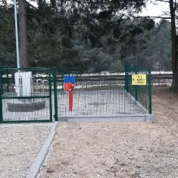 Zabezpieczona ogrodzeniem stacja wodociągowa z dwoma zaworami oznaczonymi kolorami, tabliczka informacyjna projektu na tle leśnego krajobrazu i torów kolejowych.