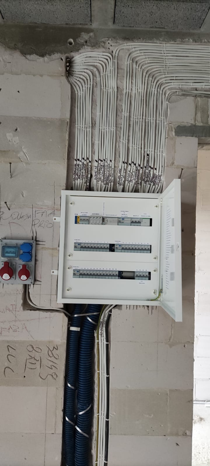 Rozdzielnica elektryczna z otwartymi drzwiczkami zamontowana na ścianie z instalacją kablową. Widoczne kable w peszlach i oznaczenia na przewodach.
