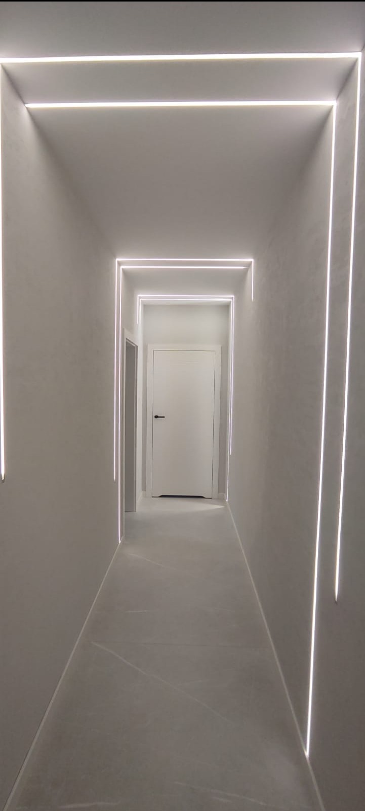 Nowoczesny korytarz z minimalistycznym oświetleniem LED tworzącym geometryczne ramy. Białe ściany i podłoga, zamknięte drzwi na końcu korytarza.