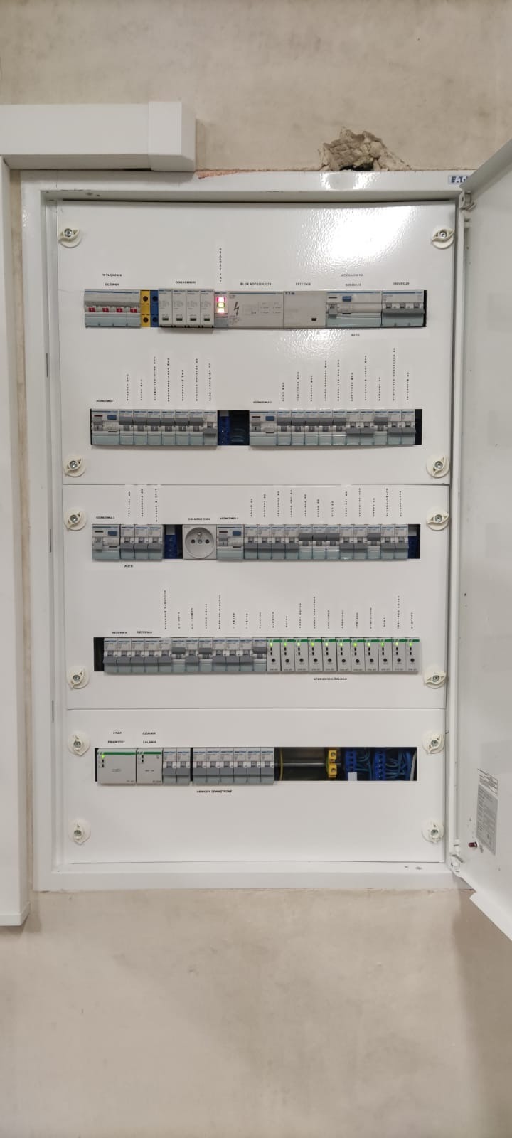 Rozdzielnica elektryczna w białej obudowie, widok z przodu. Zainstalowane zabezpieczenia, gniazdo. Detal: uszkodzenie ściany nad obudową.