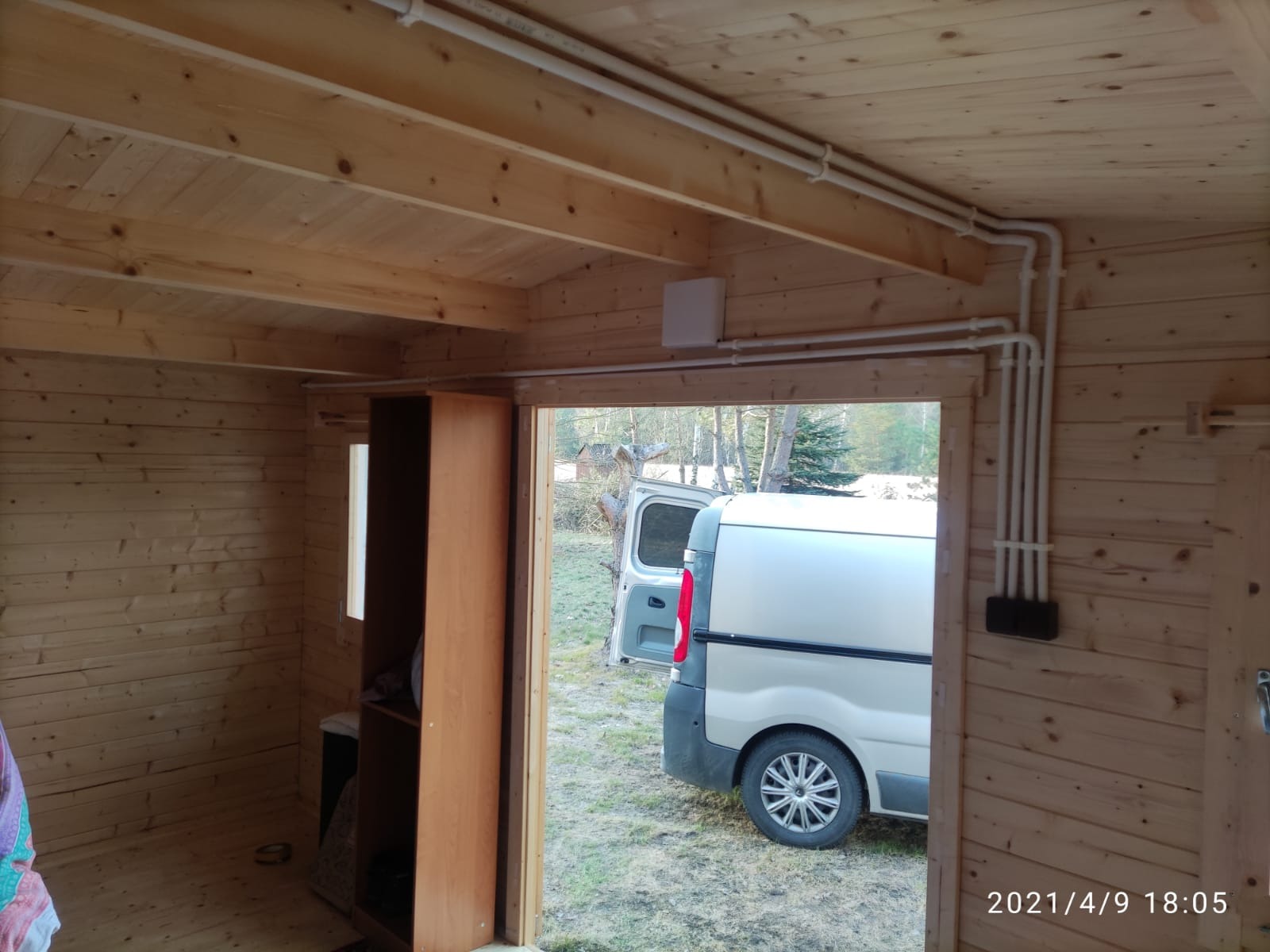 Wnętrze drewnianej altany z widoczną instalacją elektryczną w rurkach PCV, zamontowaną pod sufitem. Na zewnątrz stoi samochód dostawczy.