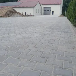 Szara kostka brukowa ułożona na dużym placu przed budynkiem z brązowym dachem i panelami słonecznymi, widoczna pryzma piasku i żwiru w tle.