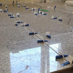 Monika Ibrahim - Szare płytki podłogowe w trakcie układania, z systemem poziomowania płytek (klipsy i kliny) oraz widocznym opakowaniem zestawu poziomującego Diall i kartonem z napisem 'Granit polerowany'.