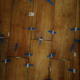 Monika Ibrahim - Układanie płytek imitujących drewno z użyciem poziomicy laserowej i klipsów poziomujących; widoczny fragment buta roboczego.