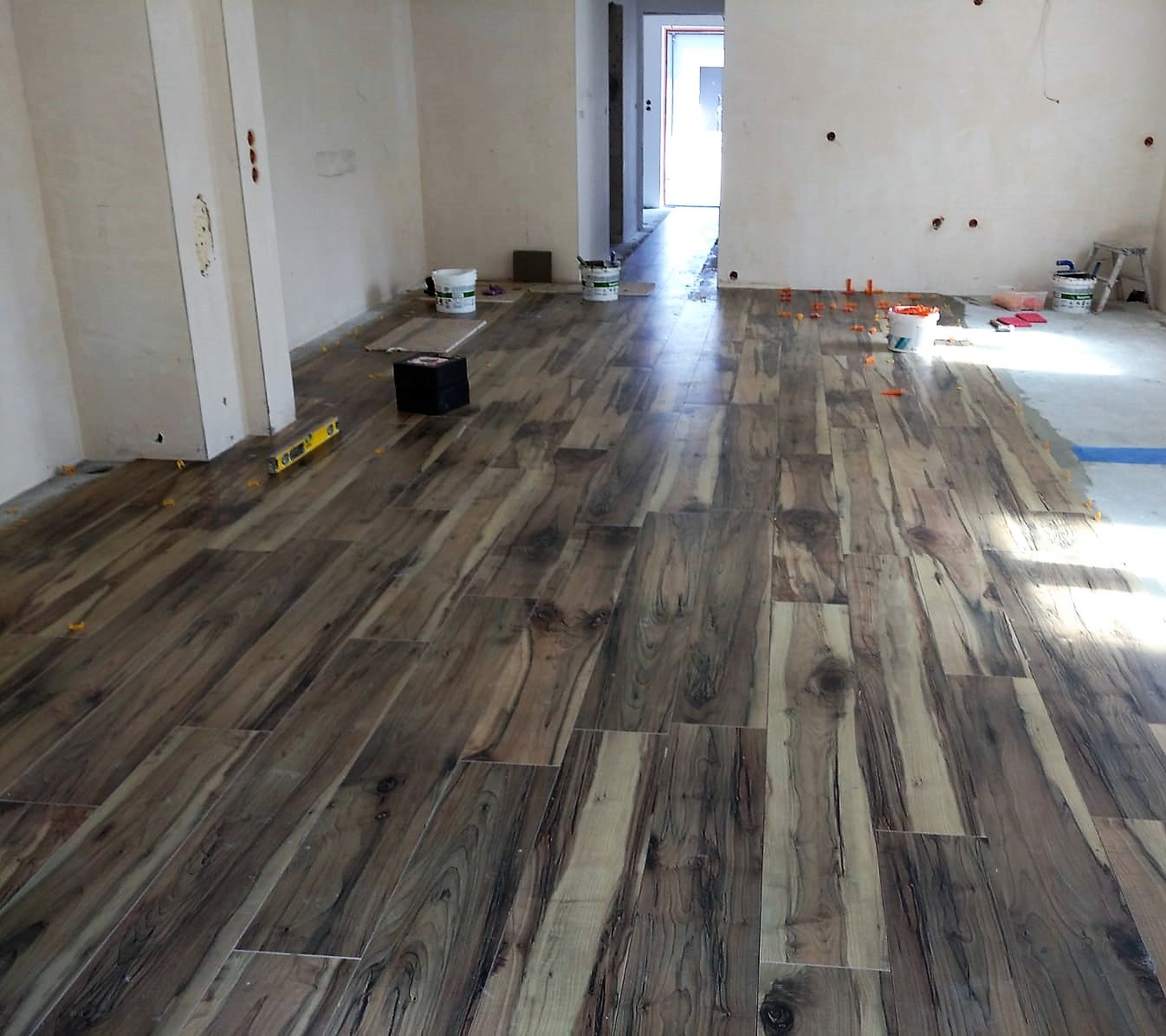 Układanie podłogi z płytek imitujących drewno w trakcie prac wykończeniowych, widoczne poziomica, pojemniki z klejem i elementy poziomujące.