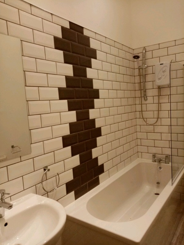Wykończona łazienka z biało-brązowymi płytkami ułożonymi w zygzak na ścianie, biała wanna i umywalka, chromowana armatura i szklany parawan nawannowy.