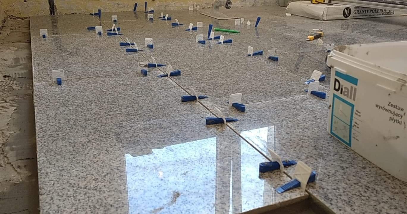 Szare płytki podłogowe w trakcie układania, z systemem poziomowania płytek (klipsy i kliny) oraz widocznym opakowaniem zestawu poziomującego Diall i kartonem z napisem 'Granit polerowany'.