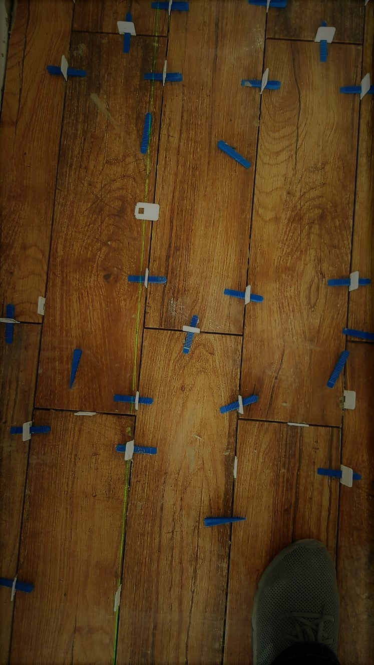 Układanie płytek imitujących drewno z użyciem poziomicy laserowej i klipsów poziomujących; widoczny fragment buta roboczego.