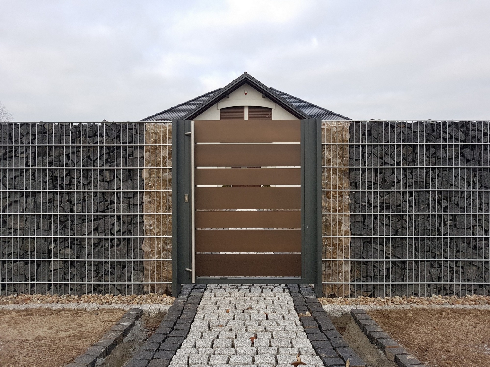 Nowoczesne ogrodzenie gabionowe z metalową bramą w Poznaniu, z widoczną ścieżką z kostki brukowej prowadzącą do wejścia.