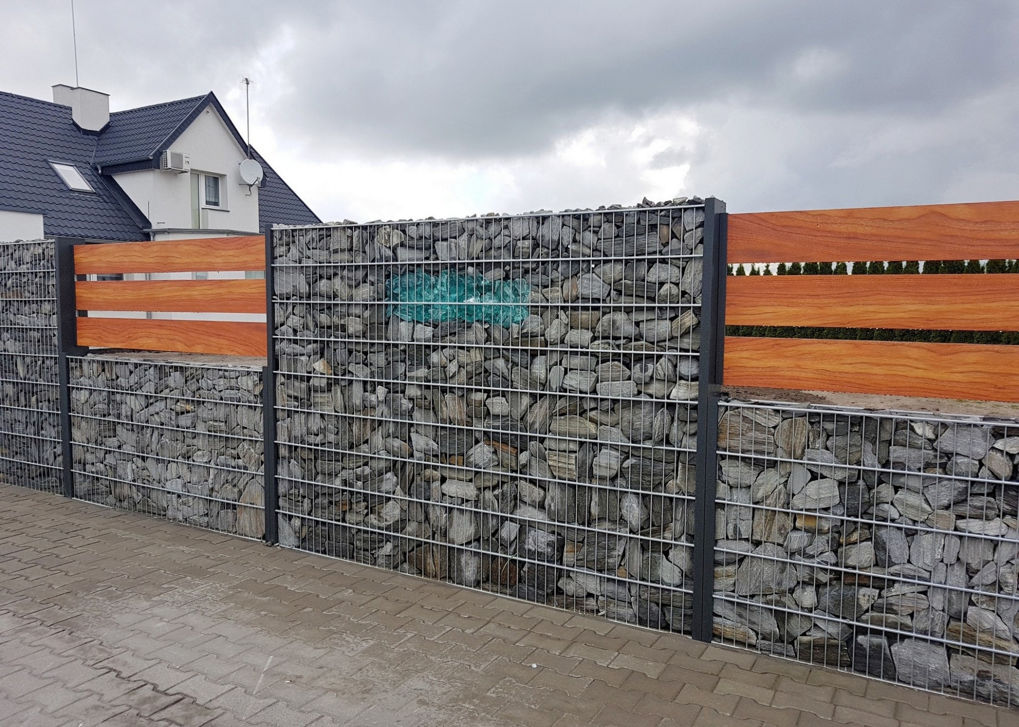 Gabionowe ogrodzenie z poziomymi elementami drewnianymi, wypełnione szarymi kamieniami, widok z zewnątrz na tle domu z ciemnym dachem i pochmurnym niebem.
