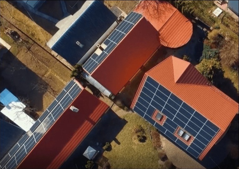 Widok z lotu ptaka na dachy budynków z panelami słonecznymi, czerwona dachówka, jesienne otoczenie.