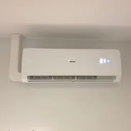 Biała klimatyzacja Haier zamontowana na ścianie w jasnym pomieszczeniu, z widocznymi przewodami w białej maskownicy i wyświetlaczem temperatury 21 stopni.