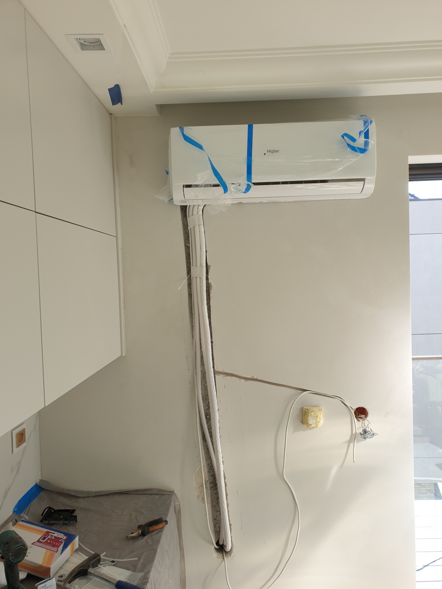 Montaż klimatyzatora Haier w pomieszczeniu, widoczne przewody w korytku, narzędzia na podłodze zabezpieczonej folią, jednostka wewnętrzna jeszcze w folii ochronnej.