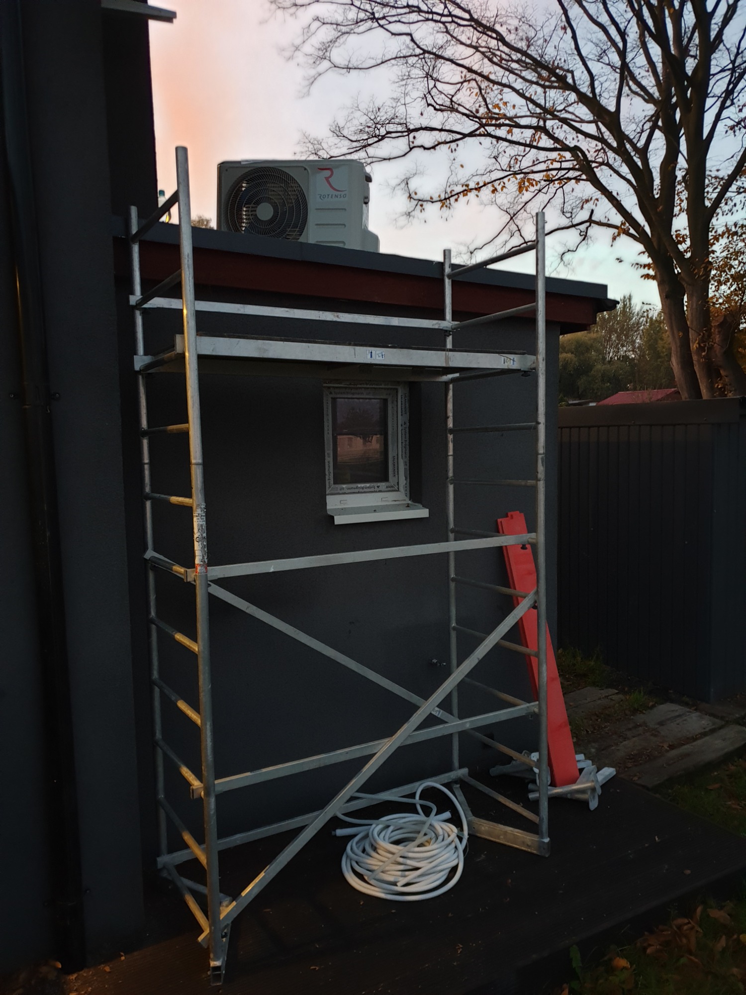 Zewnętrzna jednostka klimatyzacji marki Rotenso na dachu budynku, widoczna z poziomu rusztowania z białym zwiniętym kablem u podstawy.