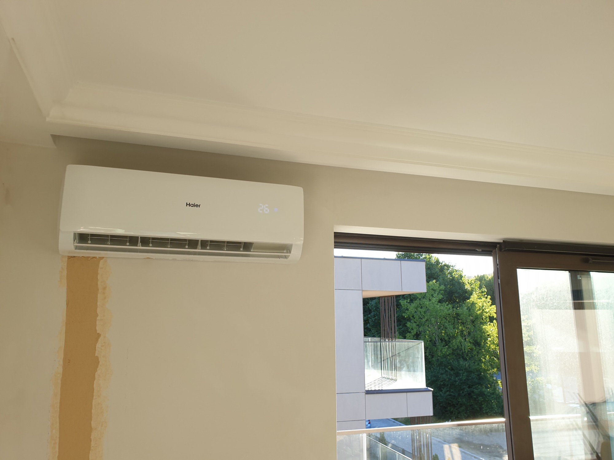 Biały klimatyzator Haier zamontowany na ścianie w jasnym pokoju, obok okna z widokiem na zieleń; widoczne ślady po montażu na ścianie.