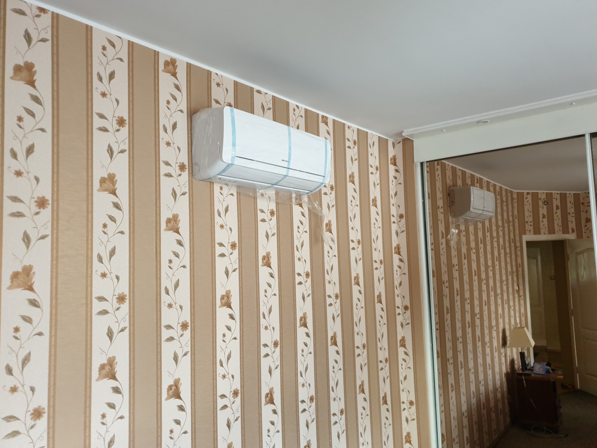Nowy klimatyzator ścienny marki Seyma, jeszcze w folii ochronnej i z taśmami, zamontowany na ścianie w pokoju z tapetą w pionowe pasy z kwiatowym wzorem. Widoczne odbicie klimatyzatora...