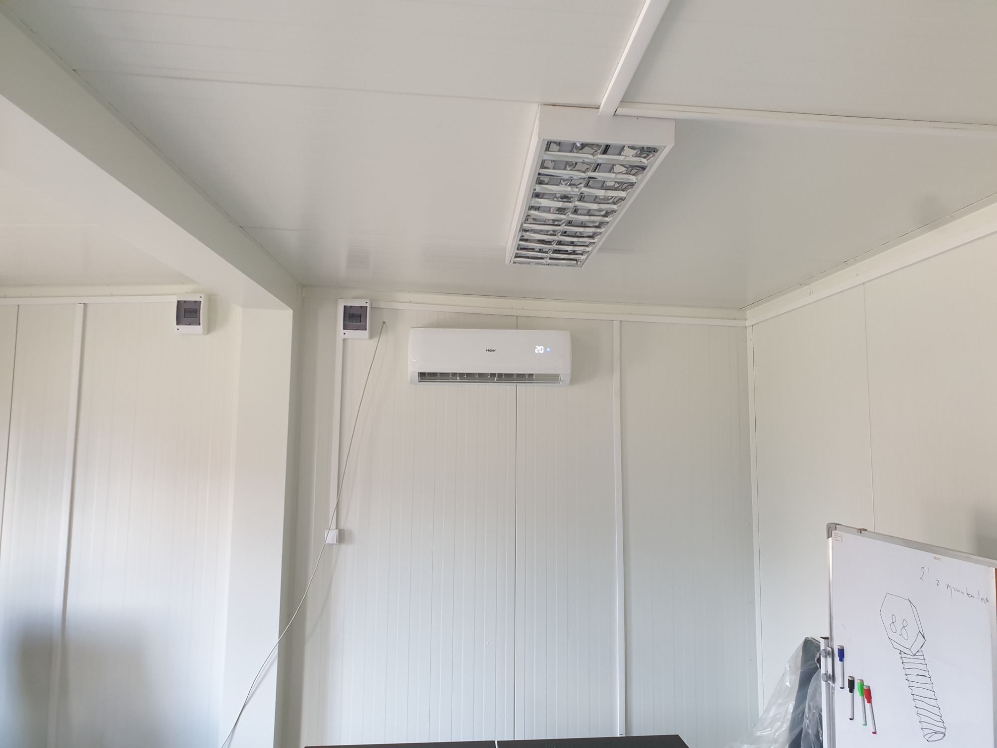 Białe wnętrze z zamontowanym klimatyzatorem Haier, widoczne skrzynki elektryczne i lampa sufitowa, częściowo widoczna tablica suchościeralna z rysunkiem śruby.