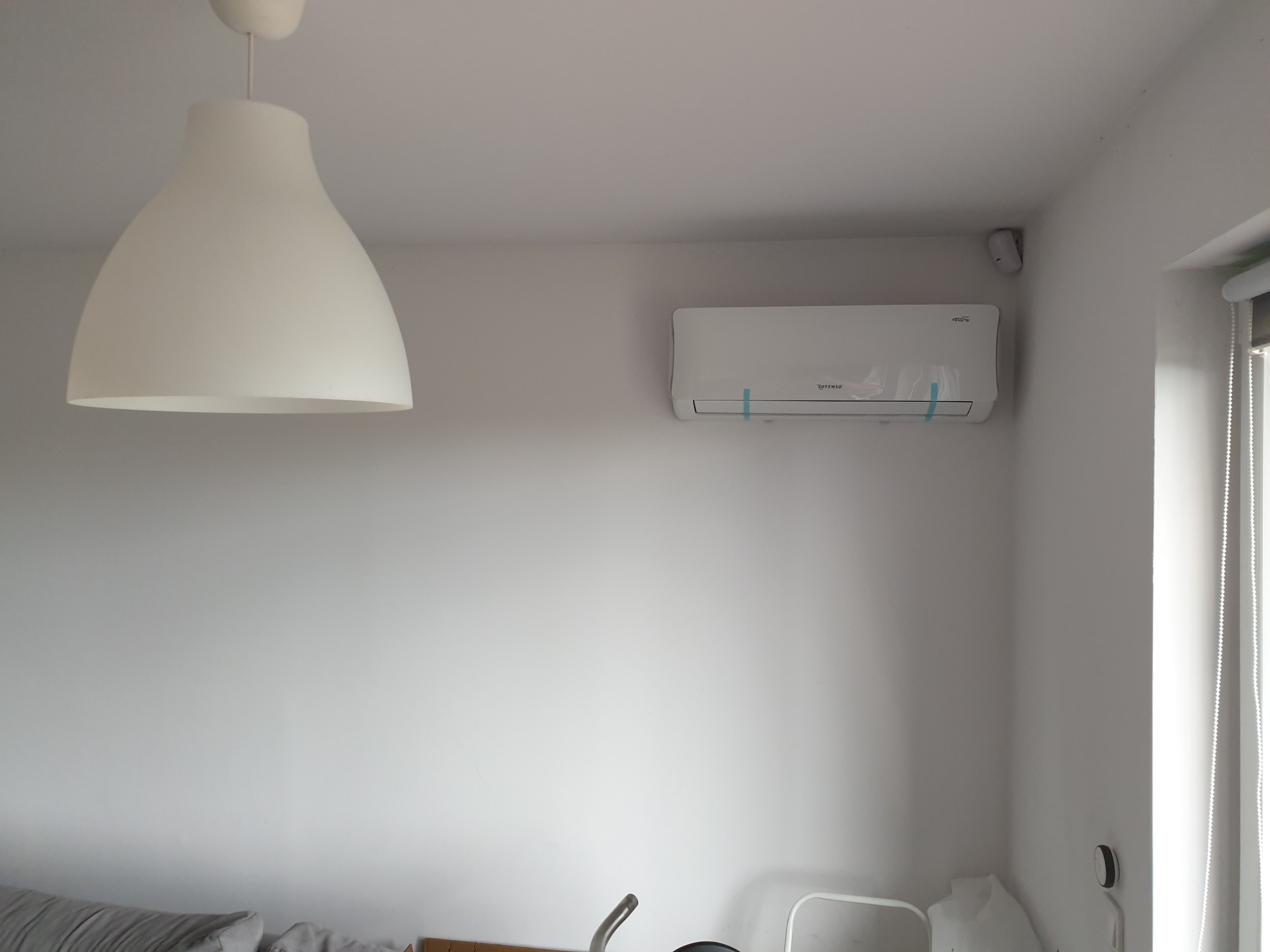 Biała klimatyzacja Rotenso zamontowana na ścianie w jasnym pomieszczeniu z widoczną lampą sufitową i roletami okiennymi, wciąż zabezpieczona folią ochronną.